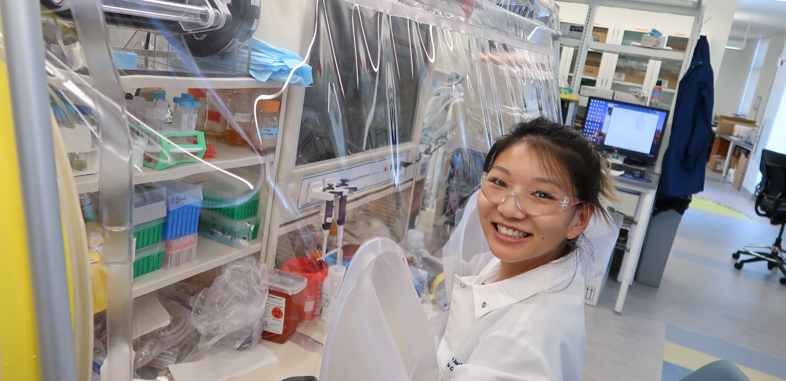 M.D./Ph.D. Candidate Jennifer Chung wearing lab coat and goggles in the Jackson Laboratory for Genomic Medicine smiling, working in an anaerobic chamber